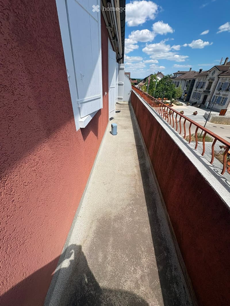Balcony with wooden railing, overlooking a town square with buildings and greenery.