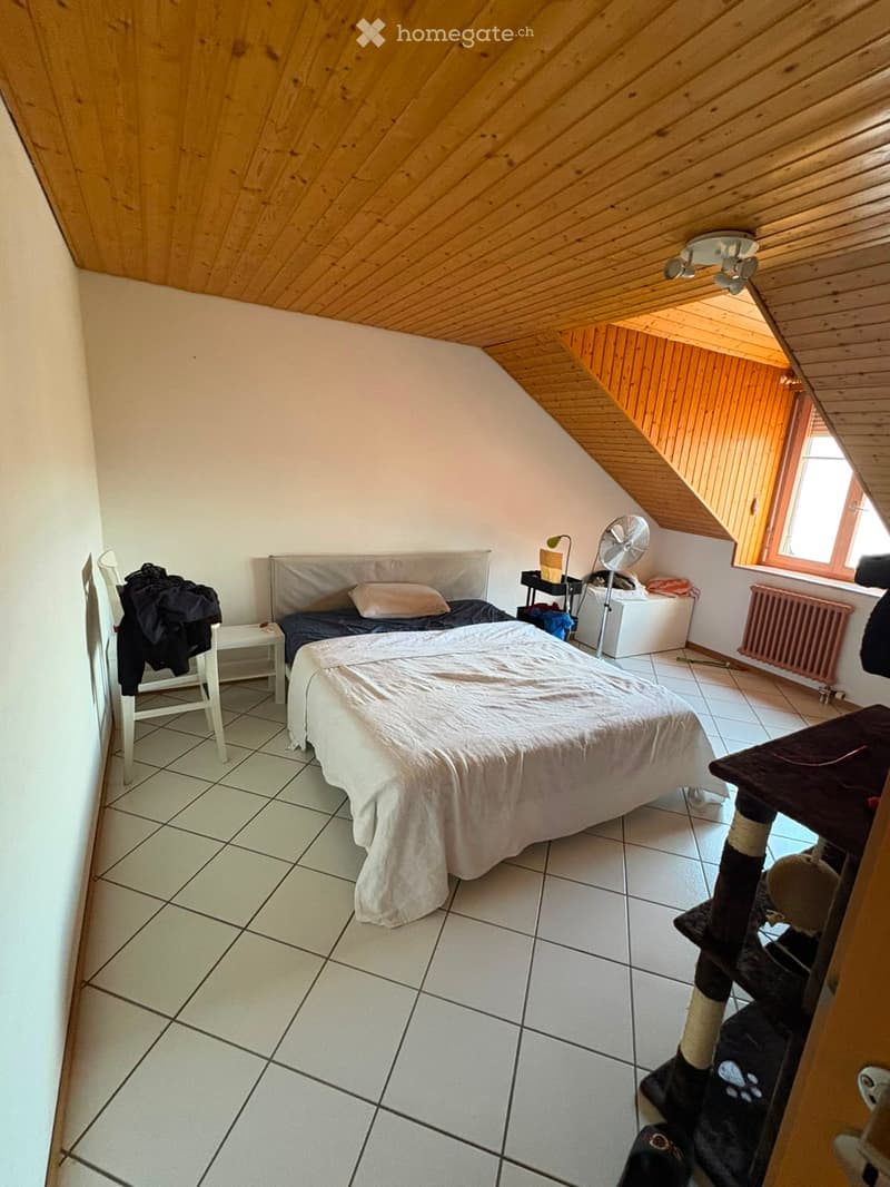 Cozy attic bedroom with wooden slanted ceilings, tiled floor, and large window. Features a double bed with white bedding, a white chair with a jacket draped over it, a cat tree, a small fan, and a radiator.