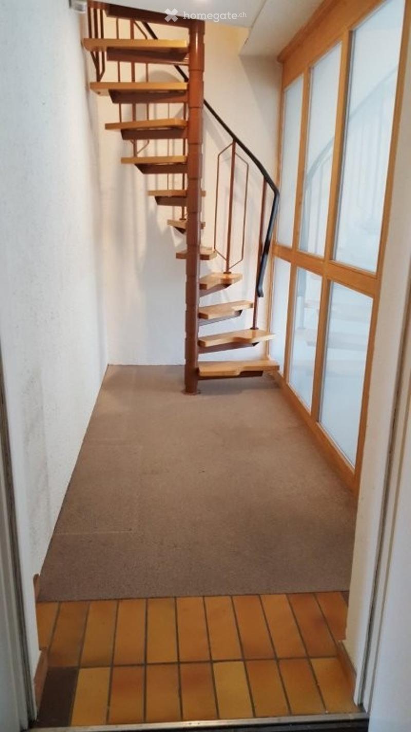 Spacious hallway with a wooden spiral staircase, beige carpet, and large windows with wooden frames.