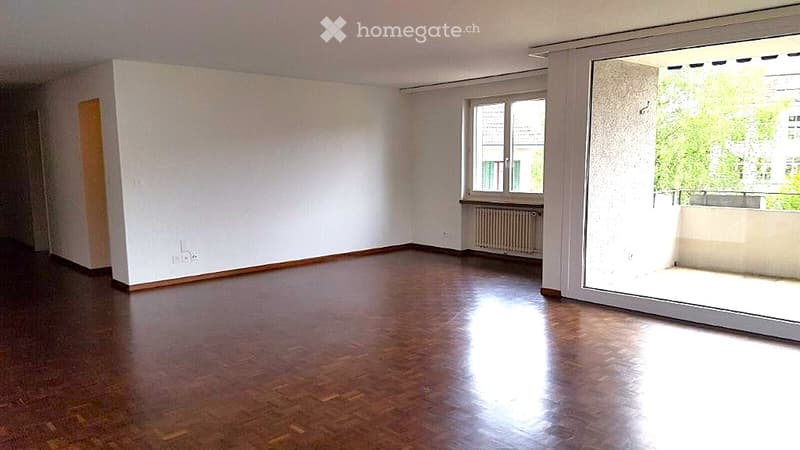 Empty room with wooden flooring, white walls, and large windows, including a sliding glass door leading to a balcony.