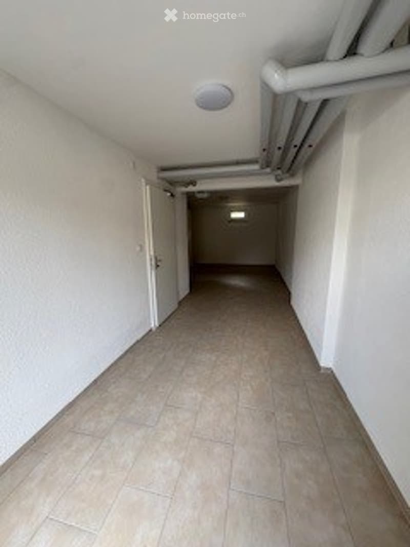 Empty hallway with tiled floor and white walls, featuring a closed door at the end and exposed ceiling pipes.