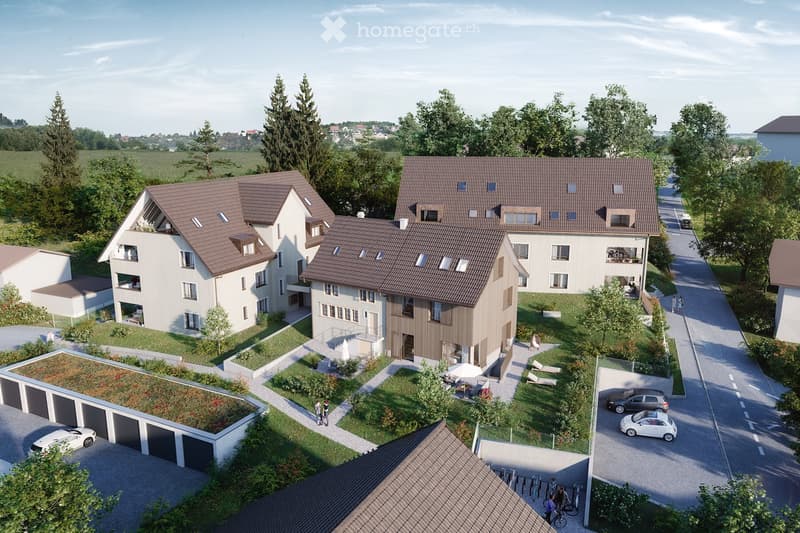 Aerial view of a residential complex with multiple modern houses, garages, and landscaped gardens. The houses feature brown tiled roofs, large windows, and balconies. The area includes a central green space with lounge chairs, a small umbrella, and a few