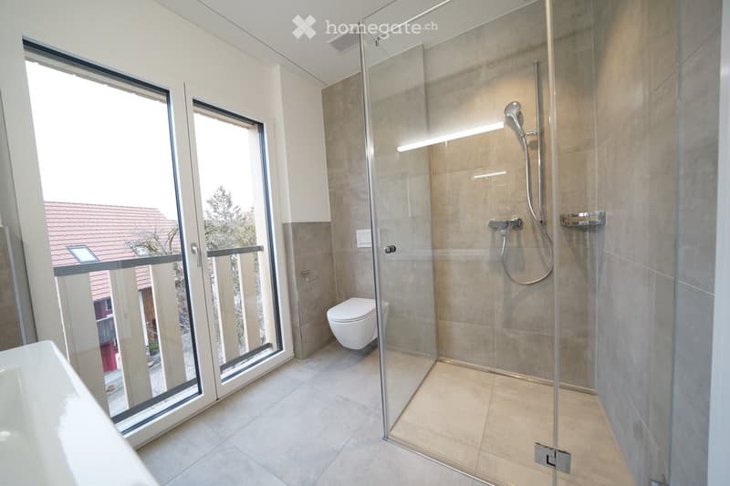 Modern bathroom with a glass-enclosed shower, toilet, and bathtub. Large windows offer a view of the outdoor area.