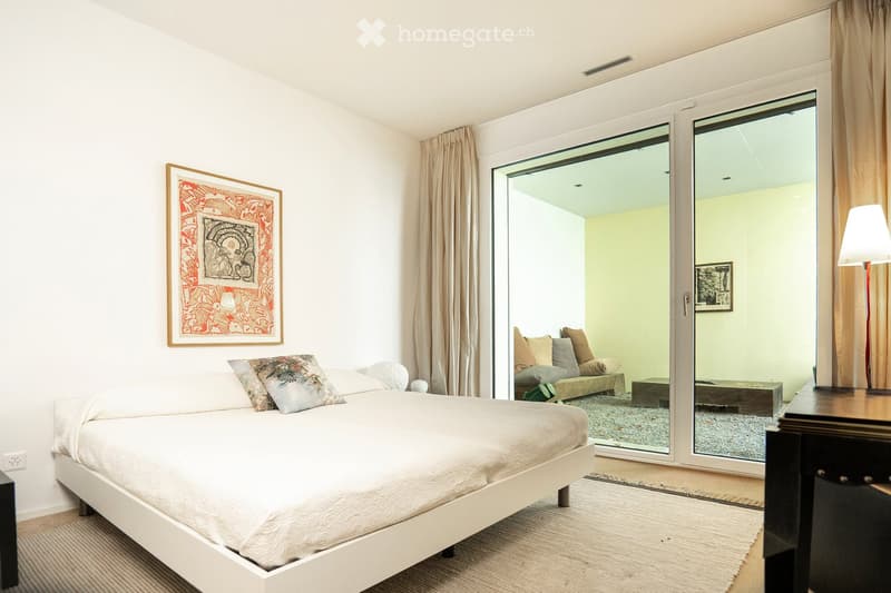 Modern bedroom with a white bed, nightstand, lamp, and artwork on the wall. Large glass doors open to a living area with a sofa and coffee table.