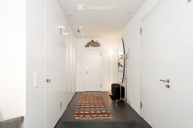 Modern, minimalist hallway with white walls, dark flooring, and a colorful runner. Features built-in storage cabinets, a coat rack, and a small bench.