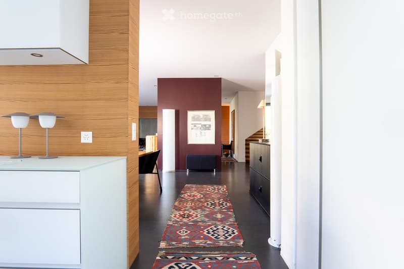 Modern hallway with dark flooring, wooden paneling on one wall, and a series of colorful rugs leading through the space. There are built-in cabinets, a small table with chairs, and modern lighting fixtures.