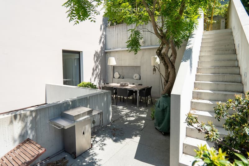 Modern outdoor patio with a dining table, chairs, a grill, and a tree providing shade. Concrete walls and steps lead to an upper level.