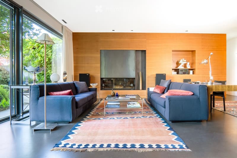 Modern living room with large windows offering a view of greenery, two blue sofas, a glass coffee table, a fireplace, and decorative items on shelves.