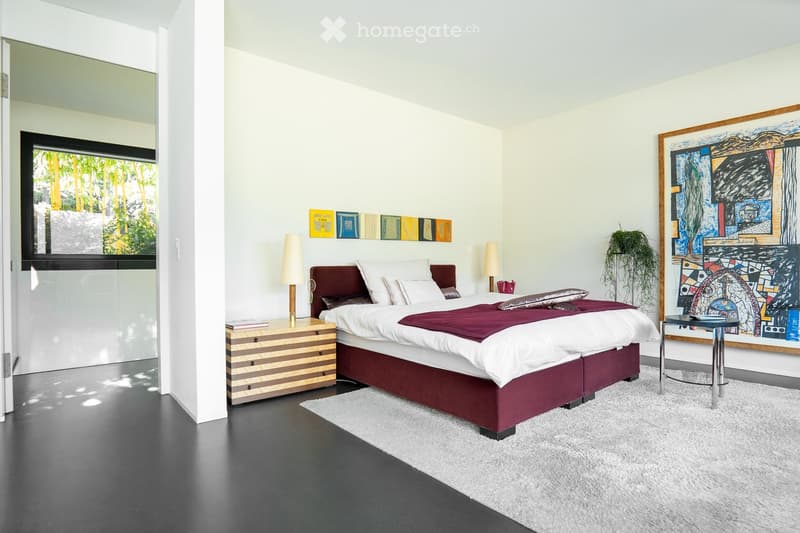 Modern bedroom with a large bed, nightstands, table lamps, a mirror, and colorful artwork on the walls. The room features a dark floor and a white shag rug.