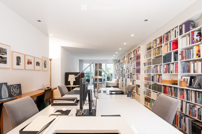 Bright, modern office space with large bookshelves, multiple desks, chairs, and a computer setup. The room features ample natural light and a sleek design.