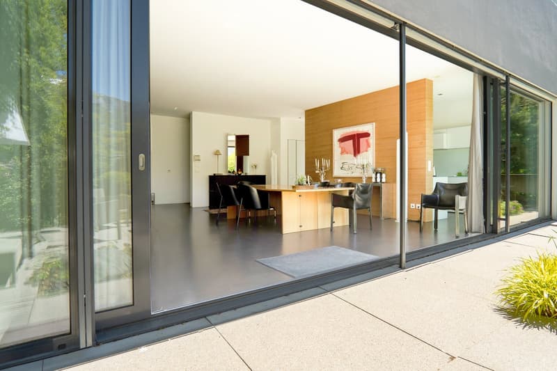 Modern, open-plan living and dining area with large glass sliding doors leading to a patio, featuring contemporary furniture including chairs, a dining table, a mirror, a lamp, and artwork on the wall.