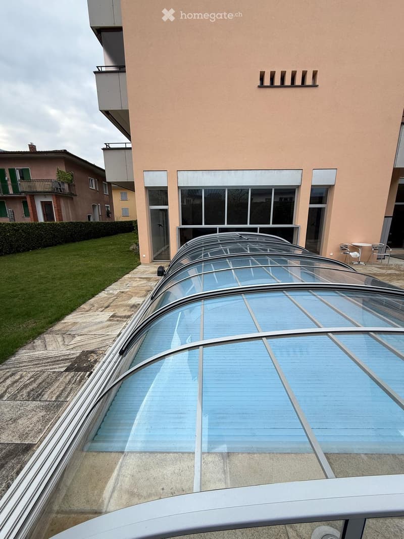 A modern glass-enclosed balcony or terrace with a curved, transparent roof structure. The area is adjacent to a building with large windows and a grassy lawn visible in the background.