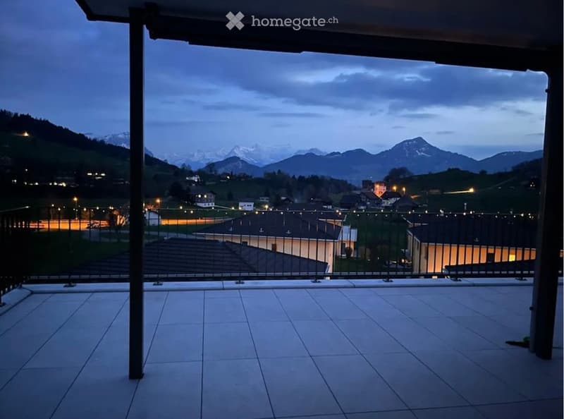 Balcony with tiled floor, metal railing, overlooking a town and mountain range at dusk.