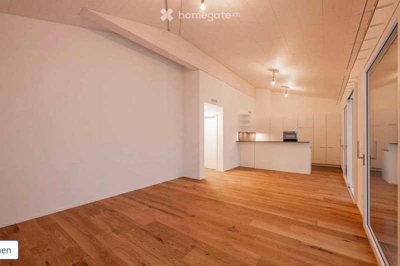 Empty room with light wooden flooring, white walls, and a doorway leading to a kitchen area with white cabinets and a countertop.