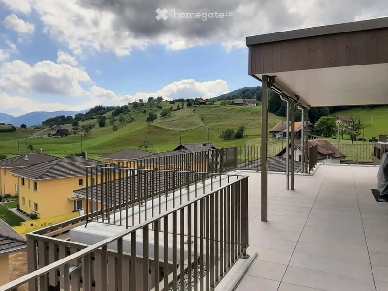 Balcony with modern metal railing, tiled floor, overlooking a green hilly landscape with scattered houses and mountains in the background.