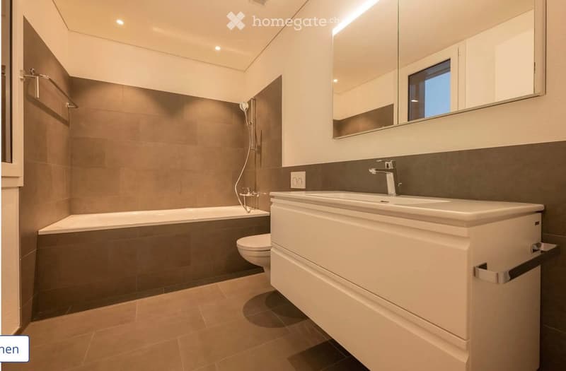 Modern bathroom with a bathtub, toilet, sink, and large mirror. Features tiled walls and a sleek design.