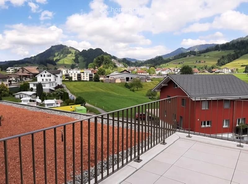 Balcony with metal railing overlooking a scenic valley, featuring houses, green fields, and distant mountains.