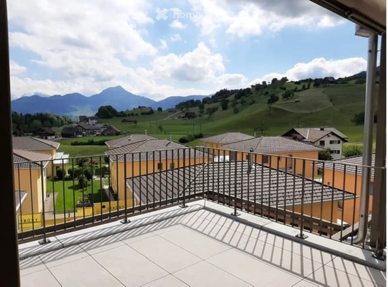 Balcony with metal railing overlooking a scenic valley and mountains, with tiled floor and clear sky above.
