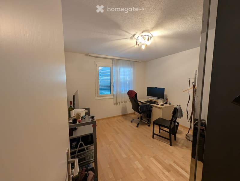 A well-lit room with wooden flooring, a desk with a computer and chair, a small shelf with various items, and a window with blinds and curtains.
