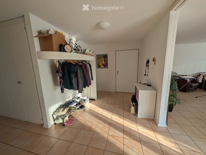 Hallway with tiled floor, coat rack, shoe rack, and a small table with a plant. There is a door leading to another room with a sofa visible.