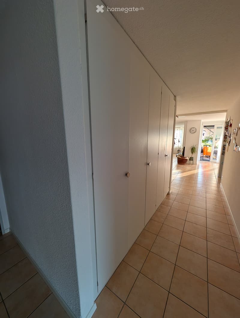 A narrow hallway with tiled flooring and white walls, leading to an open area with furniture and decor visible in the distance.