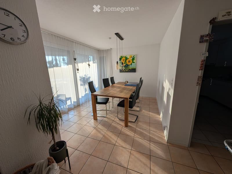 Bright dining area with a wooden dining table, six chairs, a potted plant, a wall clock, and a painting on the wall. Natural light streams in through large windows with sheer curtains.