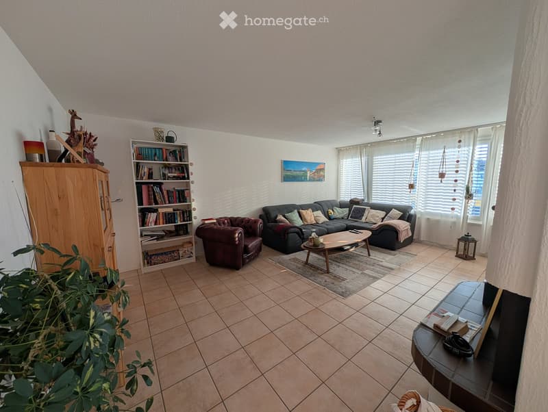 Living room with a dark grey sectional sofa, a brown leather armchair, a wooden coffee table, a bookshelf, a cabinet, and a small table with a plant. Large windows with white blinds and a painting on the wall.