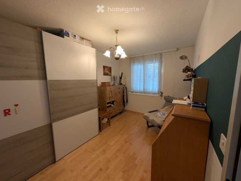 A furnished room with wooden flooring, a large mirror, a chair, a table, a cabinet, and a lamp.