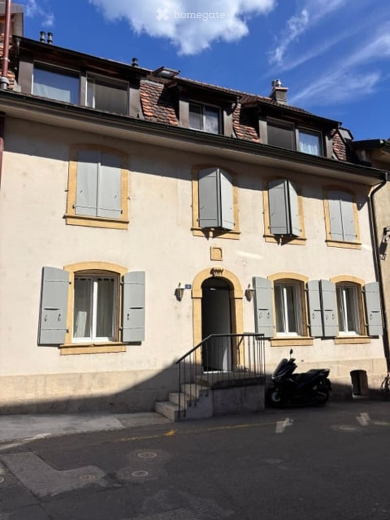 A three-story residential building with light-colored walls, multiple windows with shutters, and a small staircase leading to the entrance. A scooter is parked near the building.