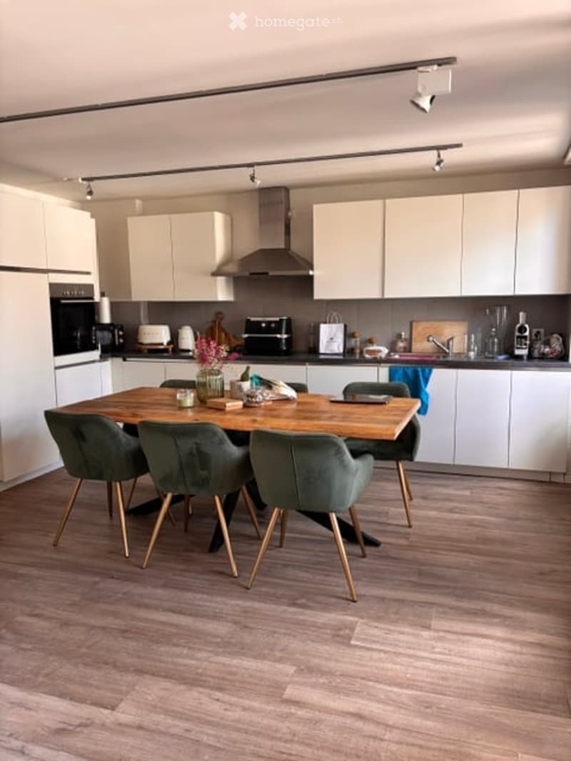 Modern kitchen with white cabinets, wooden dining table, four green chairs, and various appliances including an oven, toaster, and sink.