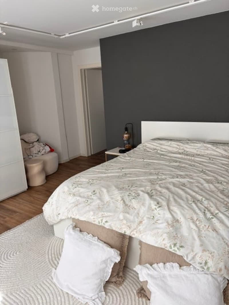 Modern bedroom with a large bed, white headboard, nightstand with lamp, wardrobe, and a small round stool near the bed.