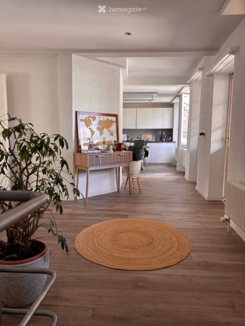 A modern, well-lit hallway with wooden flooring, a round woven rug, a small table with a plant and map, and a kitchen area visible in the background.