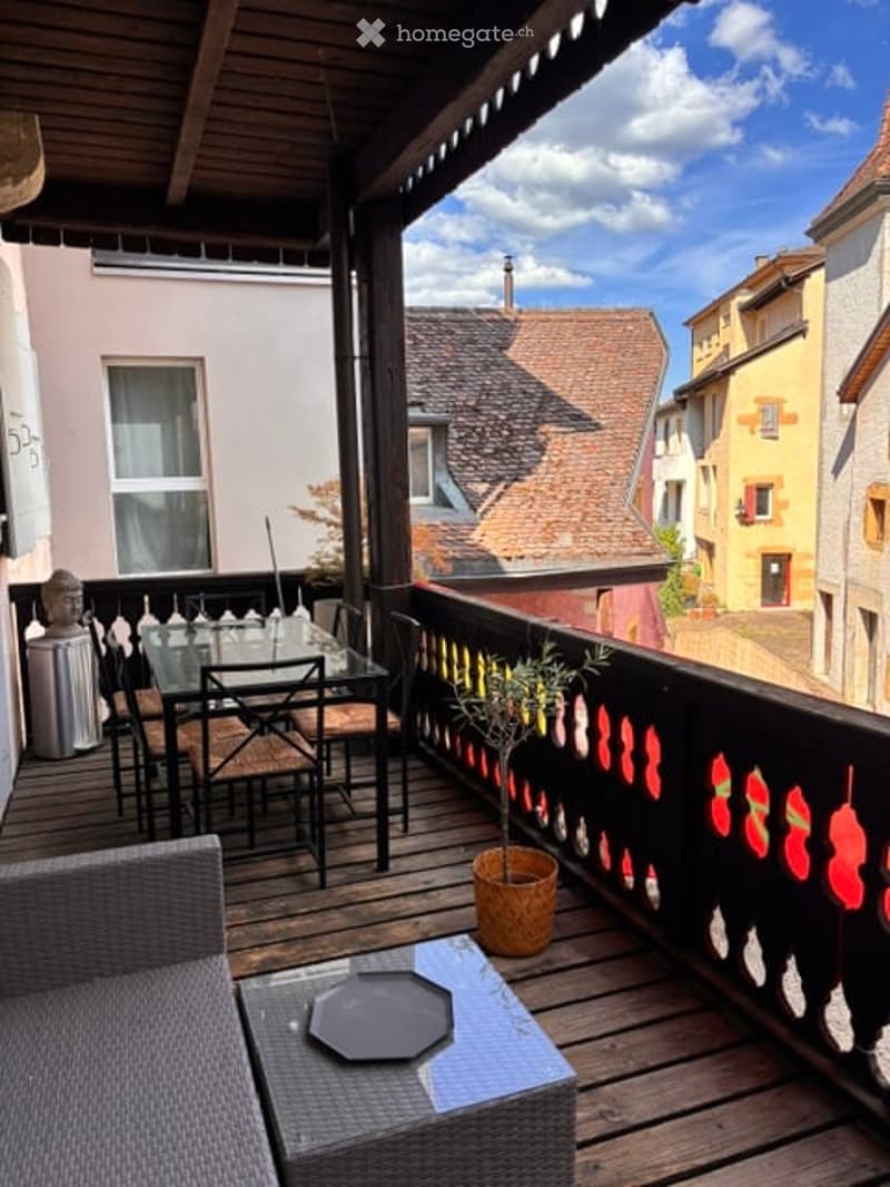 Balcony with wooden decking, outdoor furniture including a table, chairs, and a small potted plant, overlooking a historic town with colorful buildings and tiled roofs.