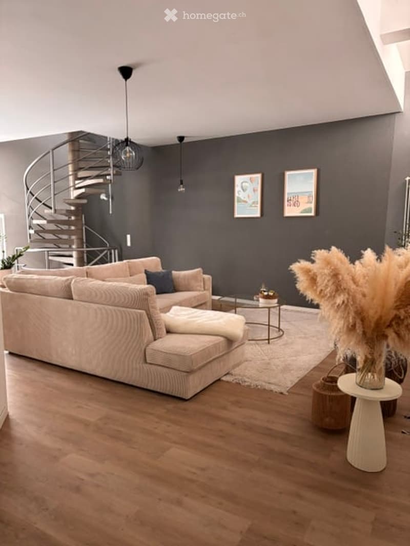 Modern living room with light wooden flooring, grey walls, a beige sectional sofa, a coffee table, a floor lamp, picture frames, a small round stool, a decorative vase with dried plants, and a woven basket.