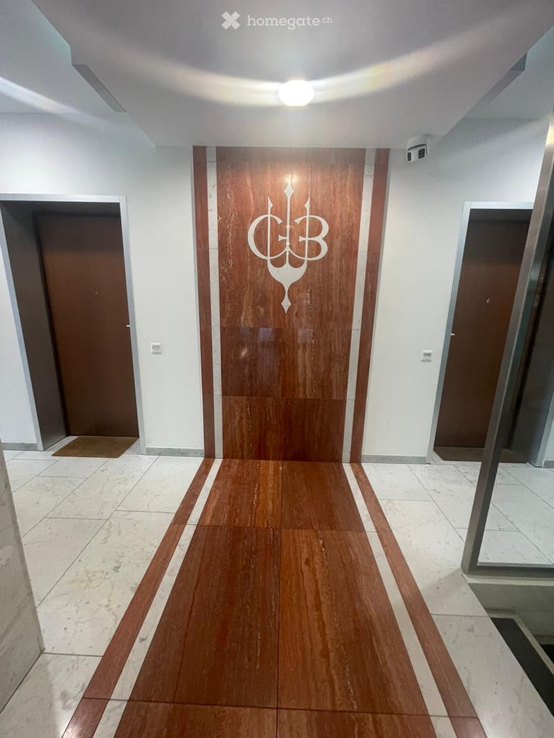 Clean, modern hallway with wooden flooring, white walls, and several doors, including a central door with a decorative logo.
