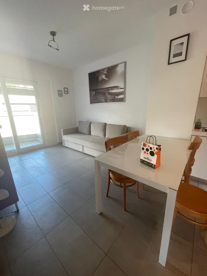Modern living room with a grey sofa, dining table with chairs, and artwork on the walls. The room has tiled flooring and large windows with a view.