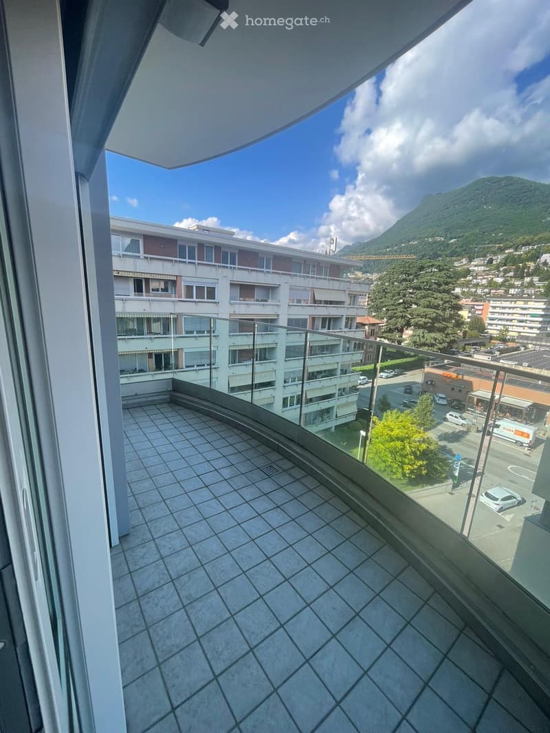 Balcony with tiled floor and glass railing, overlooking a multi-story building and a scenic mountain view.