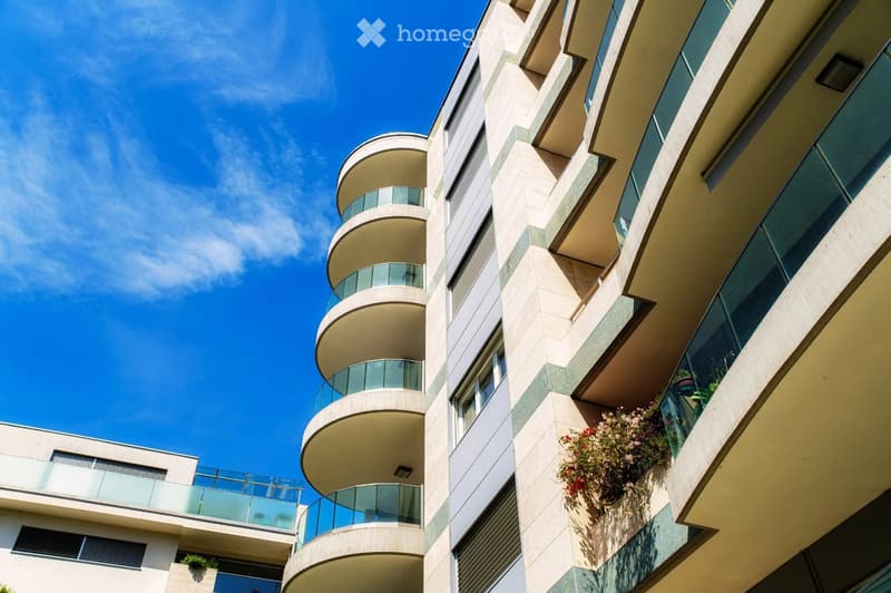 Modern multi-story residential building with curved balconies and glass railings, featuring light-colored stone facade and greenery on balconies.