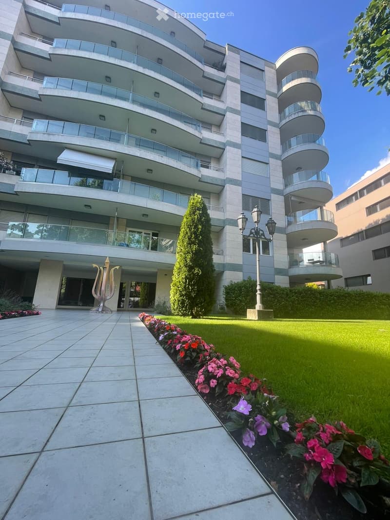 Modern multi-story residential building with balconies, surrounded by a well-maintained garden with flowers and a decorative sculpture.