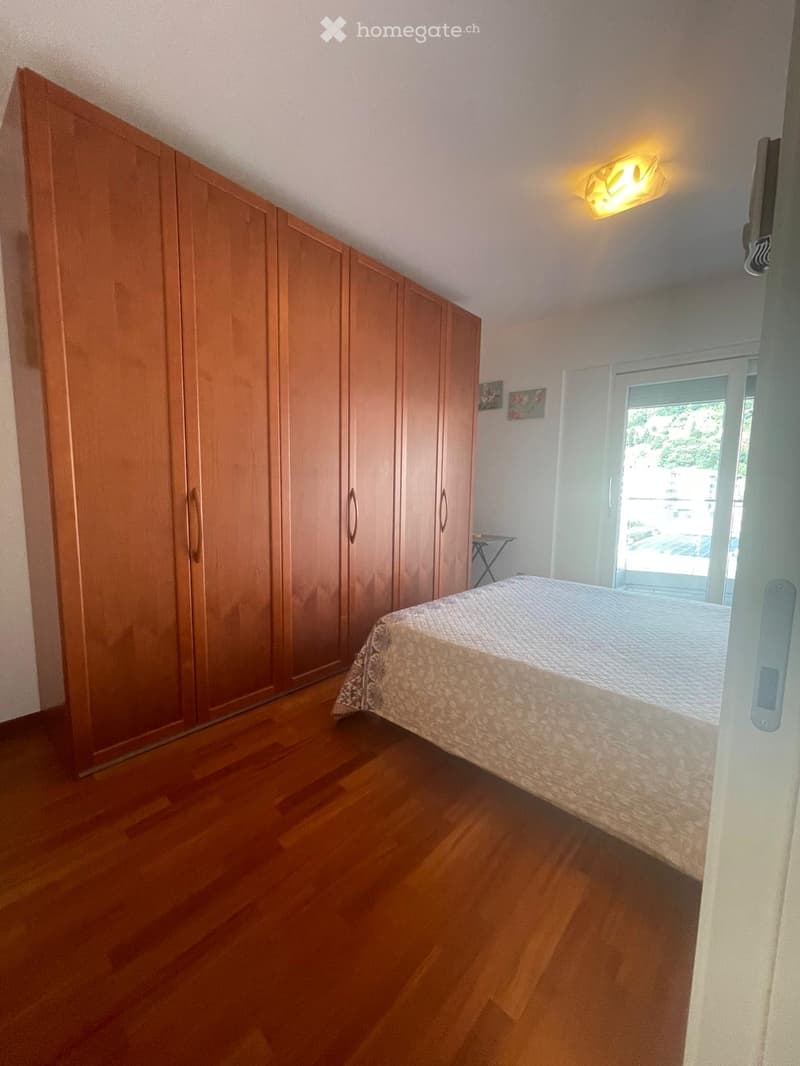 Bedroom with wooden floor, large wardrobe, bed with patterned bedspread, small table near window, and wall-mounted light fixture.