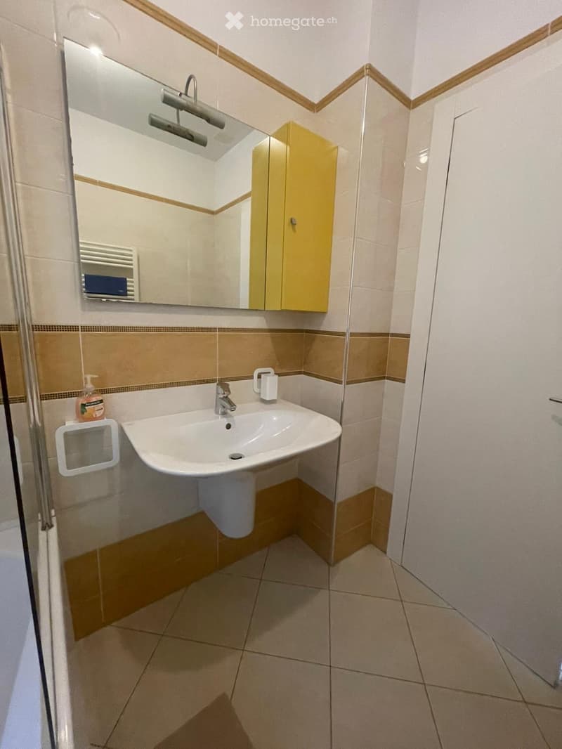 Modern bathroom with a white sink, mirror, yellow cabinet, and tiled walls.