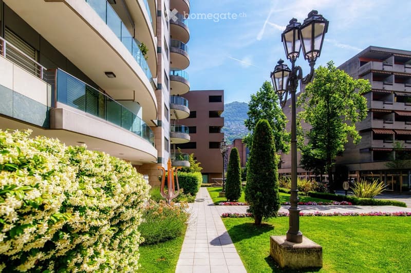 Well-maintained garden area with green lawns, trimmed hedges, flowering bushes, and a pathway leading to a modern building with balconies and large windows.