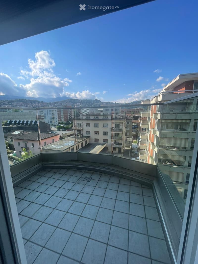 Balcony with tiled floor, glass railing, and scenic city view with buildings and mountains.