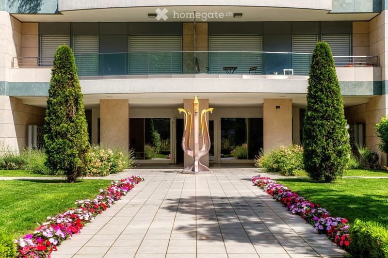 Modern building entrance with a well-maintained garden, featuring a pathway lined with colorful flowers, two tall coniferous trees, and a decorative sculpture in the center.