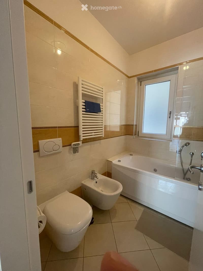 Modern bathroom with a toilet, bidet, bathtub, radiator, and a window. The walls are tiled in beige and brown, and the floor is tiled in light beige.