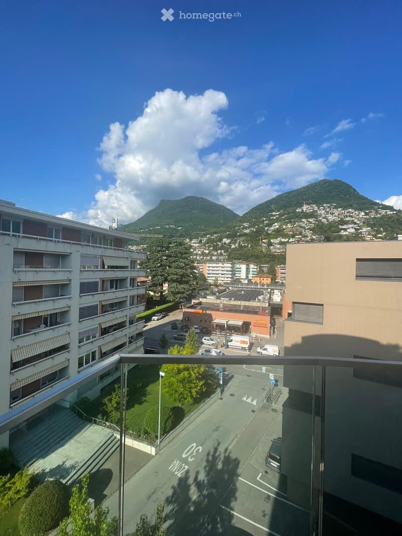 Balcony overlooking a cityscape with buildings, parking area, and distant mountains under a partly cloudy sky.