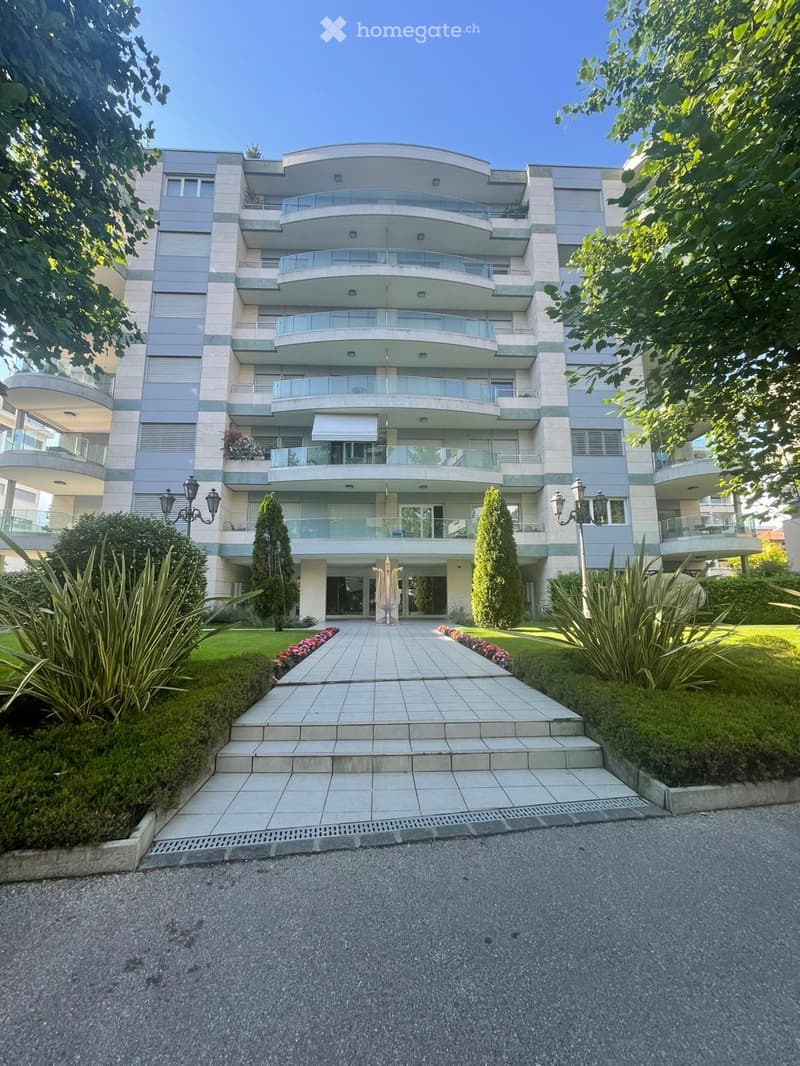 Modern multi-story residential building with balconies, surrounded by well-maintained gardens and a paved driveway.