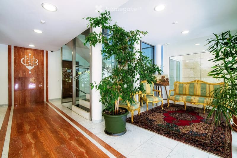 Elegant hallway with wooden flooring, large potted plants, ornate sofa and chair, and a decorative rug.