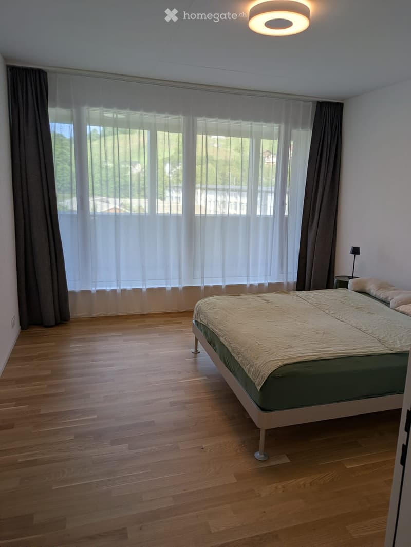 Bright bedroom with wooden flooring, large windows with sheer curtains, a double bed with a beige duvet, and a nightstand with a lamp.