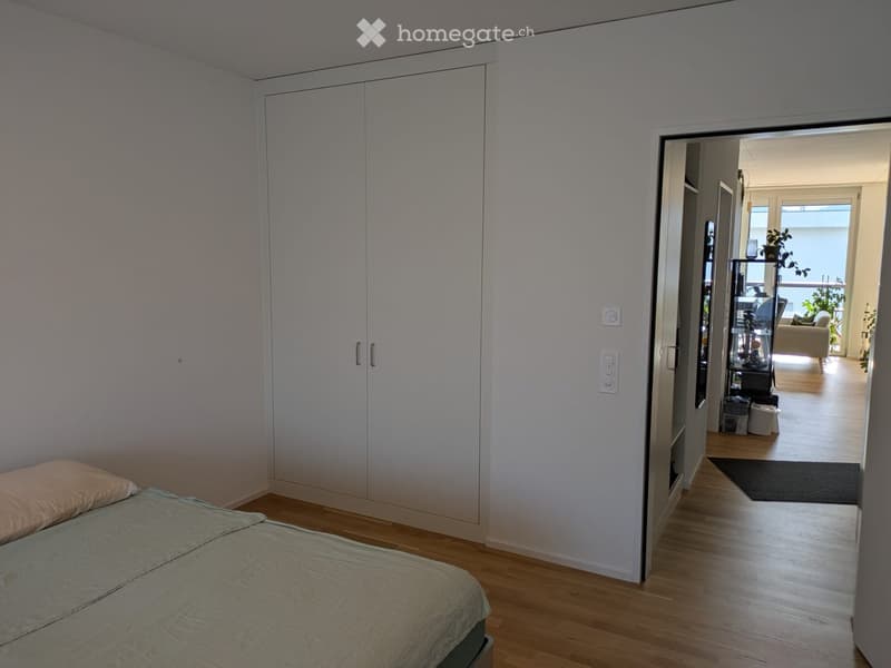 A modern bedroom with a neatly made bed, a large white wardrobe, and a view into a living area with a sofa and a balcony.